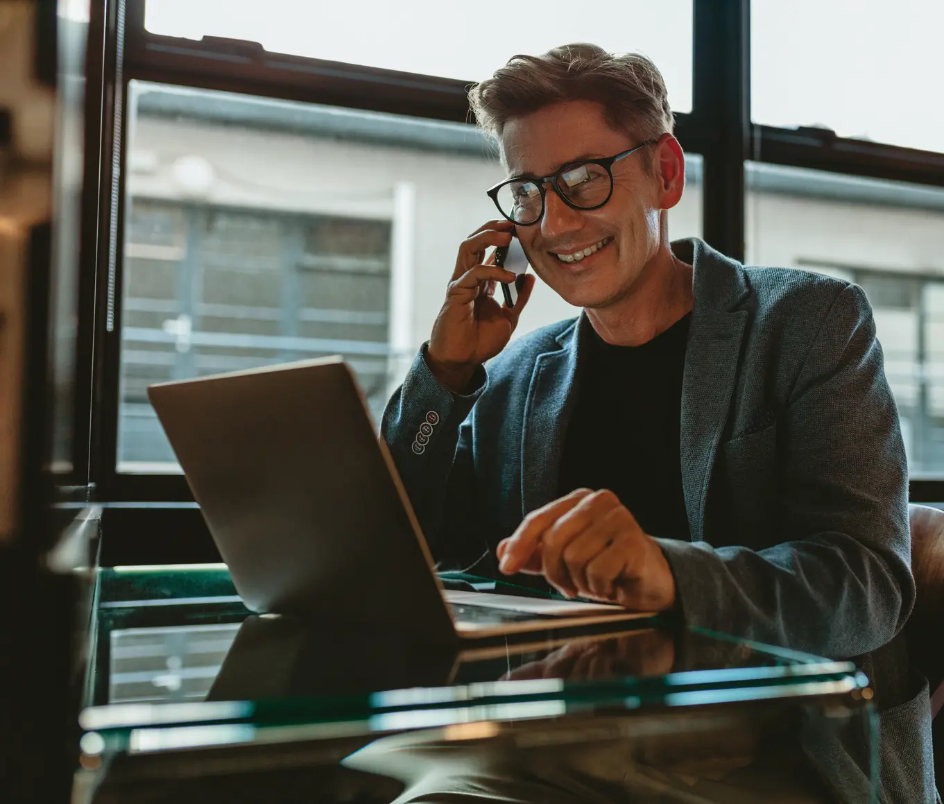 Homem de óculos, sorrindo, fala ao telefone enquanto usa o notebook perto da janela do escritório